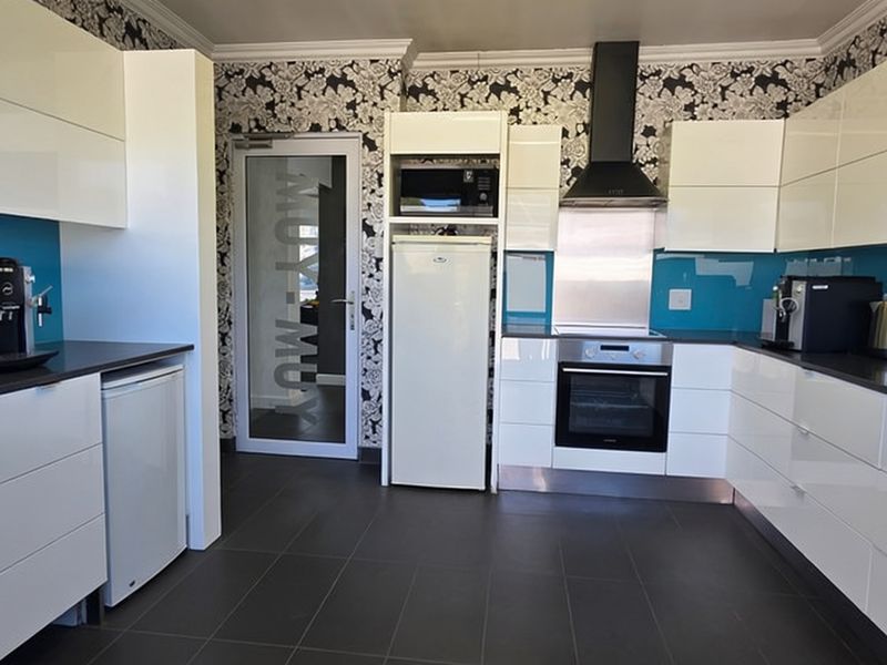 Modern office kitchen featuring high-gloss white cabinetry, blue glass splashbacks, dark floor tiles, and integrated cooking appliances.