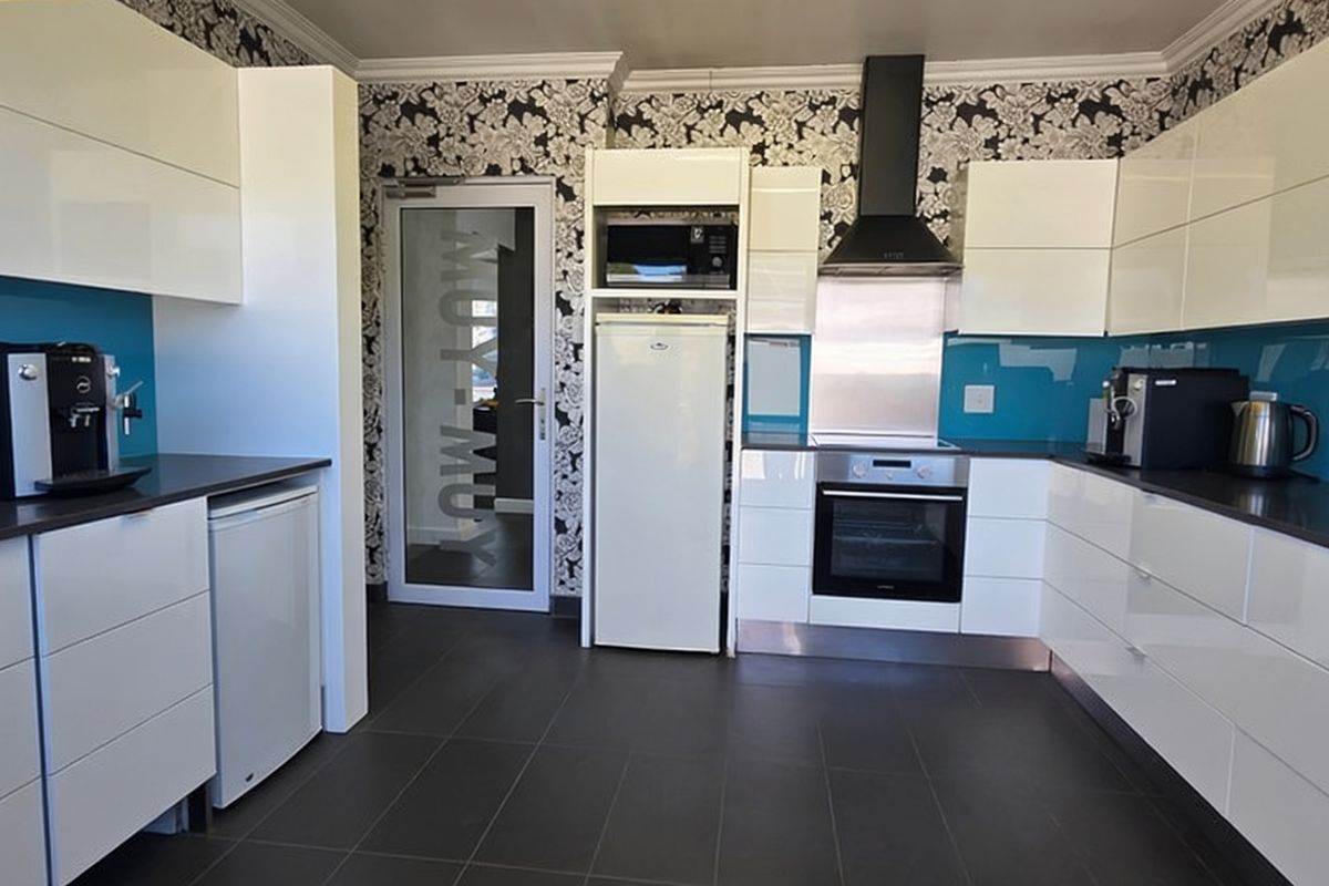 Modern office kitchen featuring high-gloss white cabinetry, blue glass splashbacks, dark floor tiles, and integrated cooking appliances.