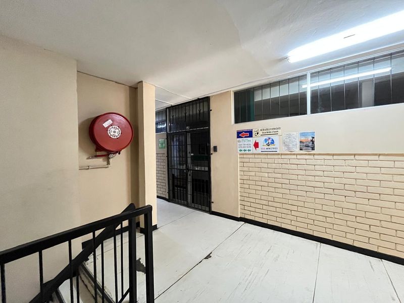 A commercial hallway featuring a fire hose reel, security gate, and brickwork wall, showing signs of wear on the flooring and walls.