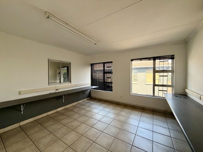 A functional, empty commercial office space featuring tiled flooring, built-in desk workstations, and large windows with venetian blinds.