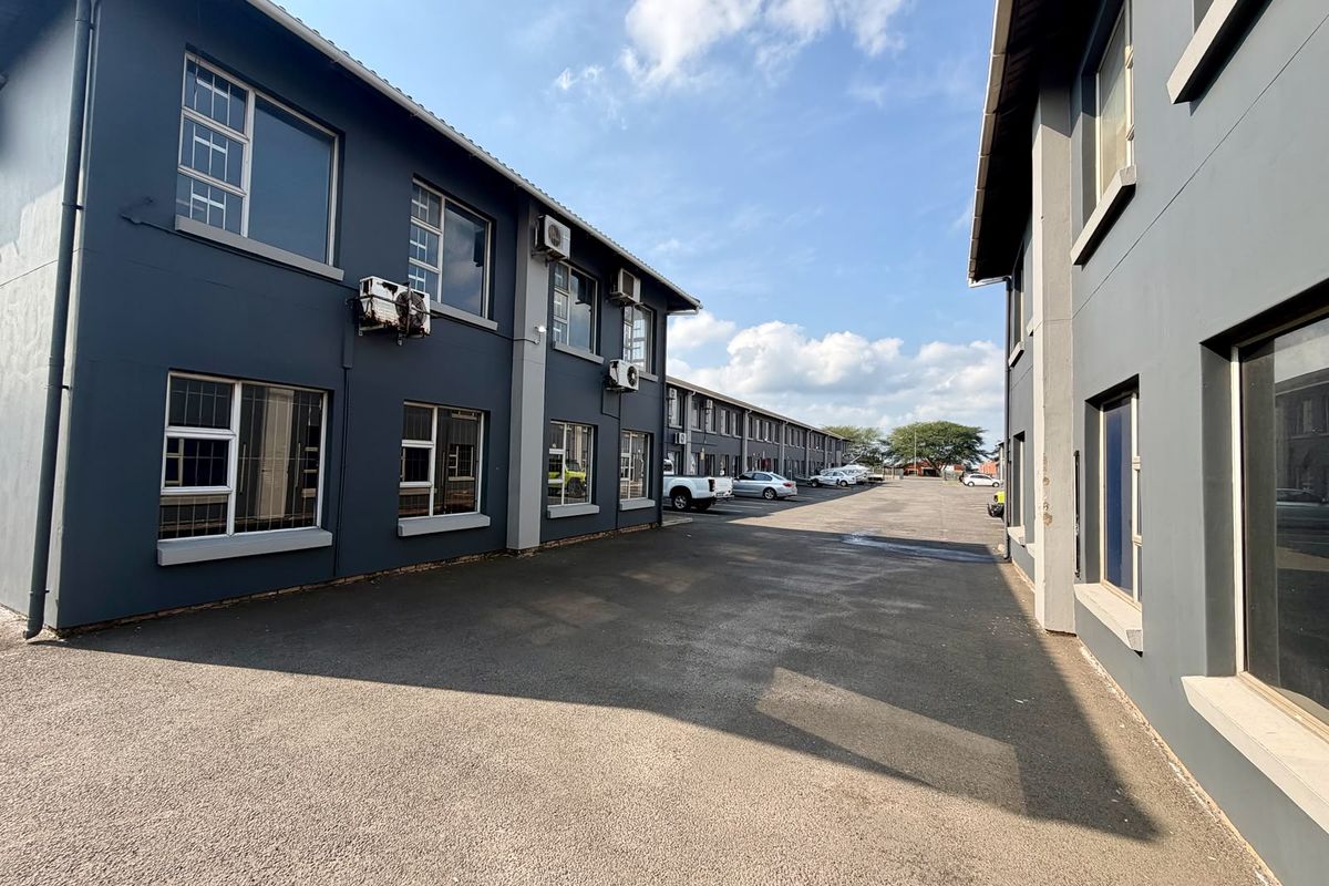 Exterior view of a commercial property complex featuring paved driveway access between two multi-story buildings with grey painted facades and aluminium window frames.