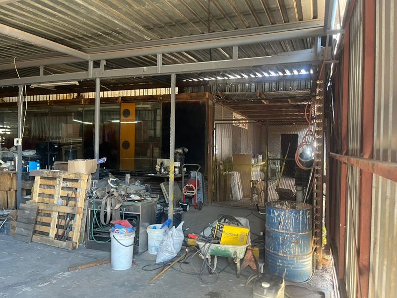 Industrial utility space featuring corrugated metal roofing, steel support beams, and a secure metal mesh gate, suitable for workshop or storage use.