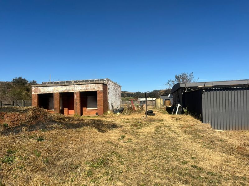 Exterior view of an industrial property featuring a brick structure and metal-clad building set on a large, unmaintained plot of land.