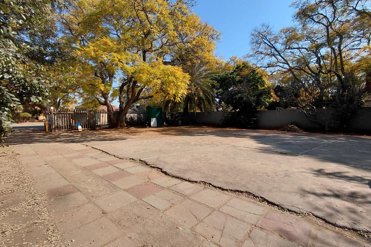 A spacious outdoor paved area featuring mature trees, perimeter walling, and a metal security gate, suitable for parking or commercial utility use.