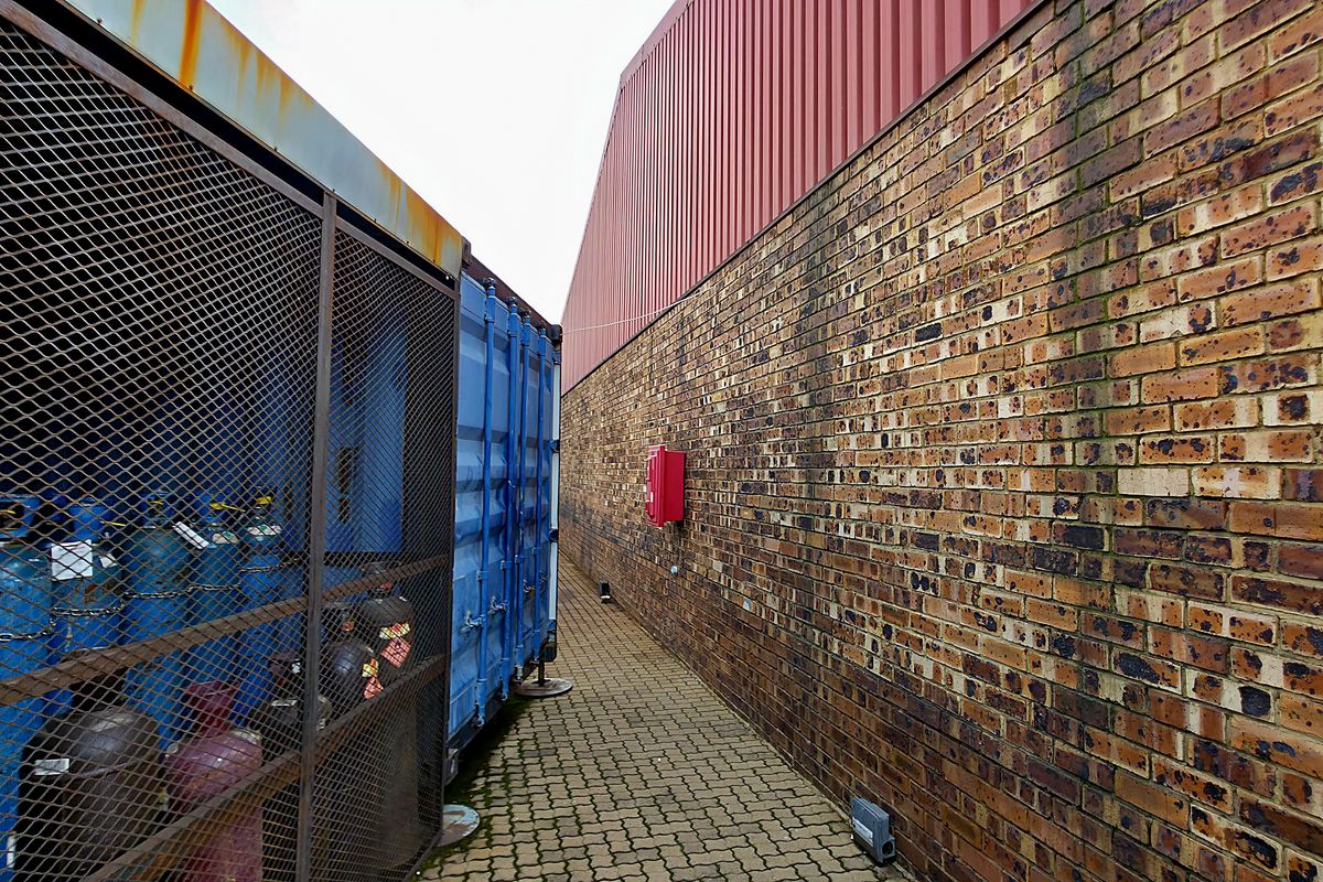 A narrow exterior service corridor featuring a brick wall, corrugated metal cladding, and a secure metal mesh storage enclosure for industrial gas cylinders.