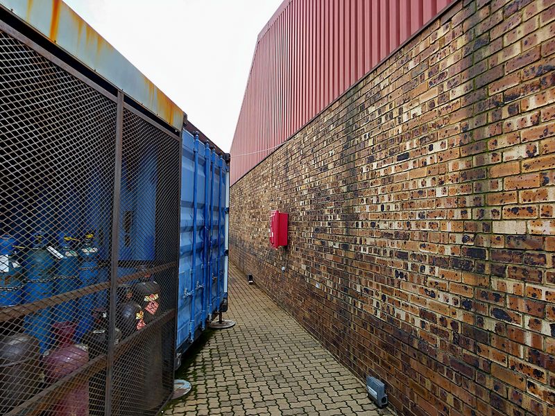 A narrow exterior service corridor featuring a brick wall, corrugated metal cladding, and a secure metal mesh storage enclosure for industrial gas cylinders.