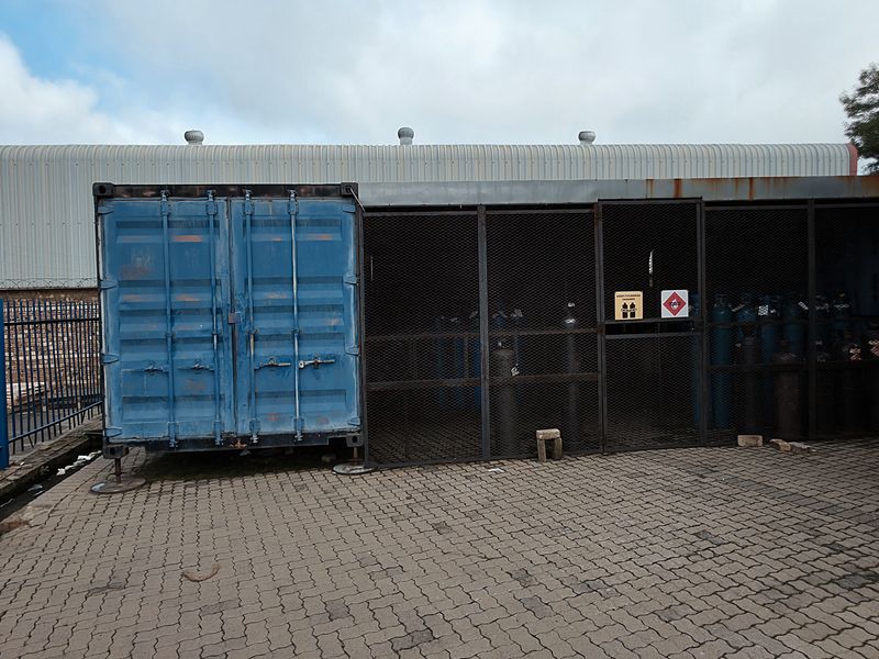 Industrial utility area featuring a secure metal storage container and mesh-enclosed gas cylinder storage unit on a paved yard.