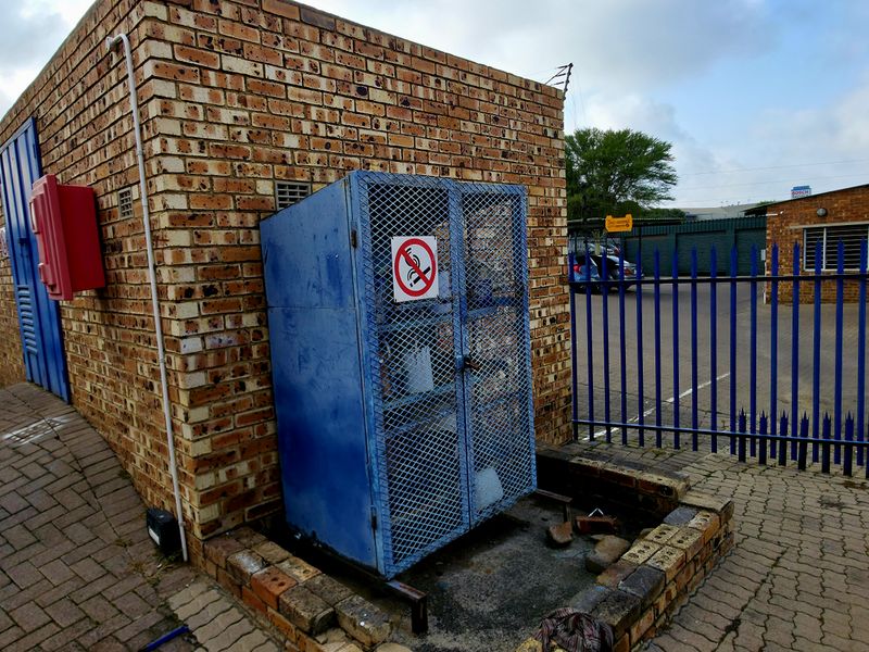 Exterior utility area featuring a face brick structure with a metal security cage and perimeter fencing on a paved industrial site.