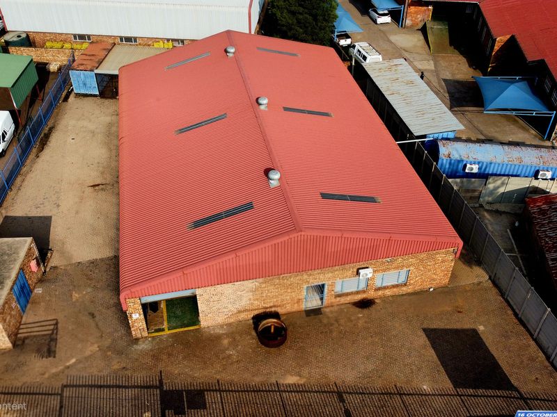 Aerial view of an industrial property featuring a large warehouse with a red corrugated metal roof, brick exterior, and surrounding paved yard area.
