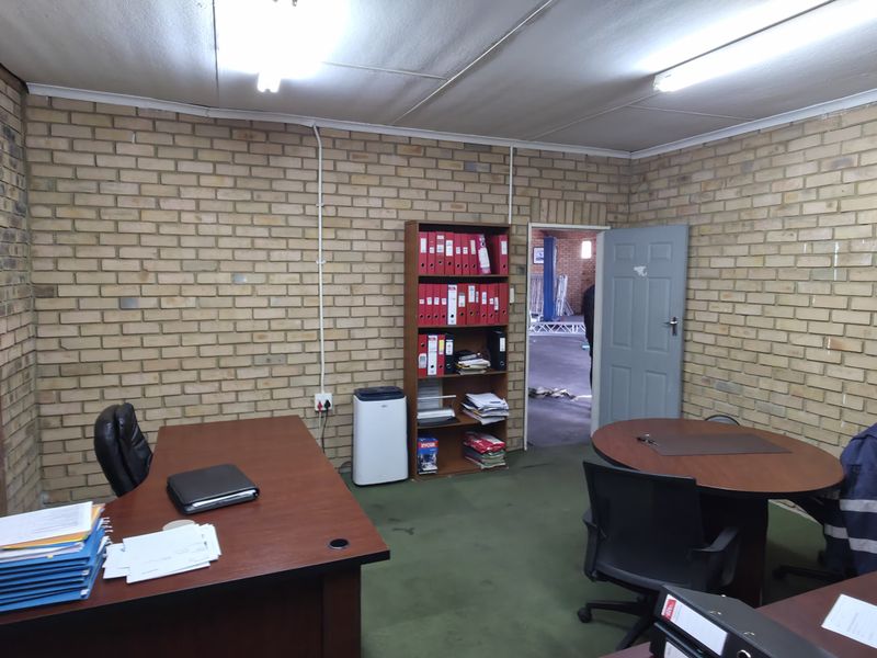 Functional industrial office space featuring exposed brick walls, fluorescent lighting, and carpeted flooring, suitable for administrative or management use.