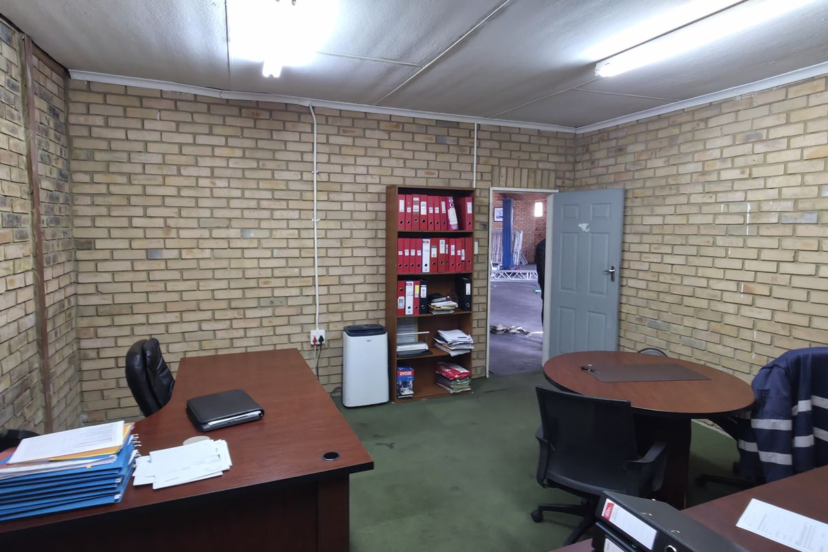 Functional industrial office space featuring exposed brick walls, fluorescent lighting, and carpeted flooring, suitable for administrative or management use.