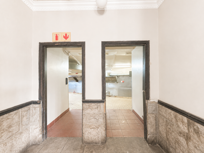 A hallway featuring stone wainscoting and crown molding, providing access to a commercial kitchen area with tiled flooring and visible ventilation infrastructure.