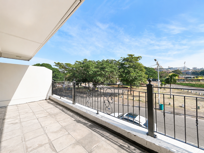 A spacious covered balcony featuring tiled flooring and a wrought iron railing, offering an open view of the surrounding area.