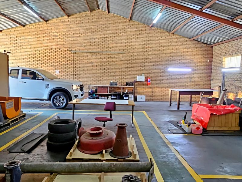 Spacious industrial utility workshop featuring exposed brick walls, steel truss roofing, fluorescent lighting, and durable painted concrete flooring with safety demarcations.