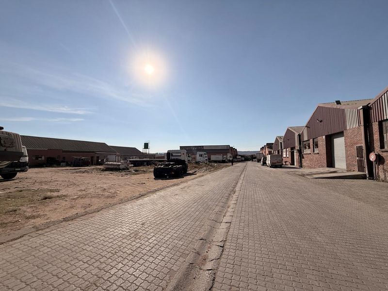 An industrial commercial complex featuring paved access roads, brick warehouse structures with roller shutter doors, and open yard space.