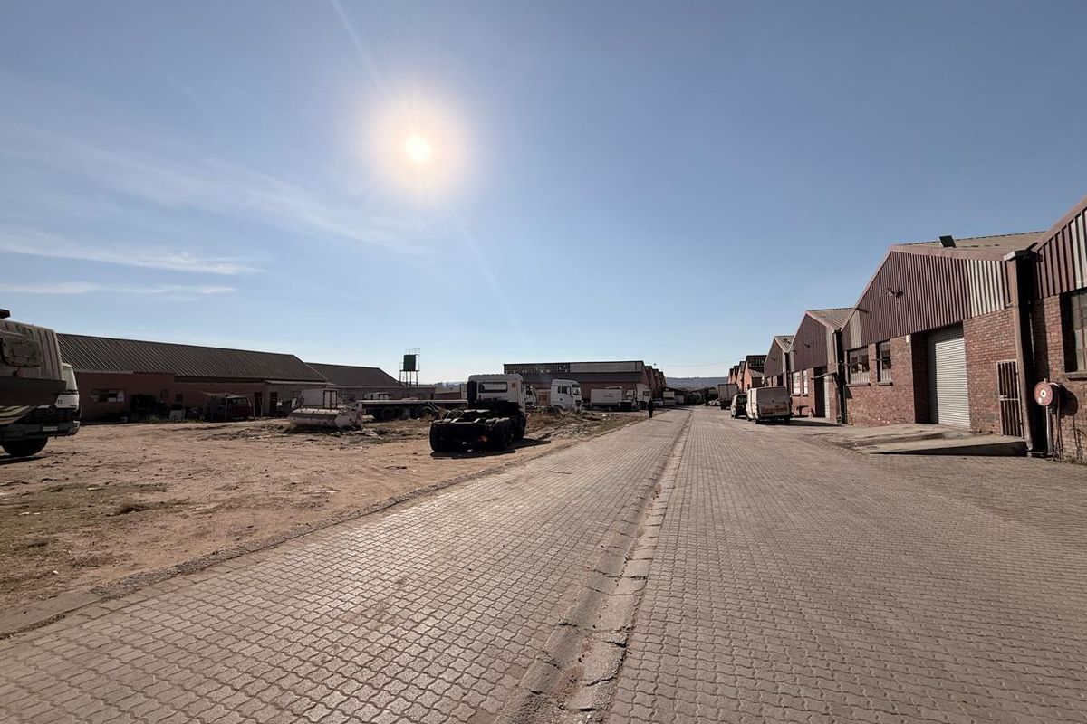 An industrial commercial complex featuring paved access roads, brick warehouse structures with roller shutter doors, and open yard space.