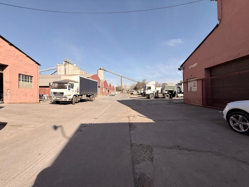 Industrial yard area featuring concrete paving, warehouse buildings, and silo infrastructure with conveyor systems, suitable for logistics or manufacturing operations.