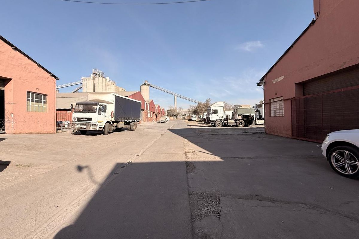 Industrial yard area featuring concrete paving, warehouse buildings, and silo infrastructure with conveyor systems, suitable for logistics or manufacturing operations.