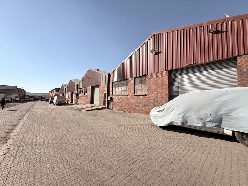 Exterior view of a commercial warehouse complex featuring brick facades, corrugated metal roofing, and multiple roller shutter doors with paved access.