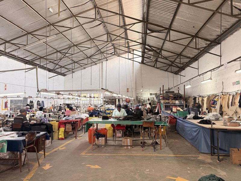 Large industrial warehouse space featuring high exposed steel roof trusses, concrete flooring, and functional lighting, currently utilized as a garment manufacturing facility.