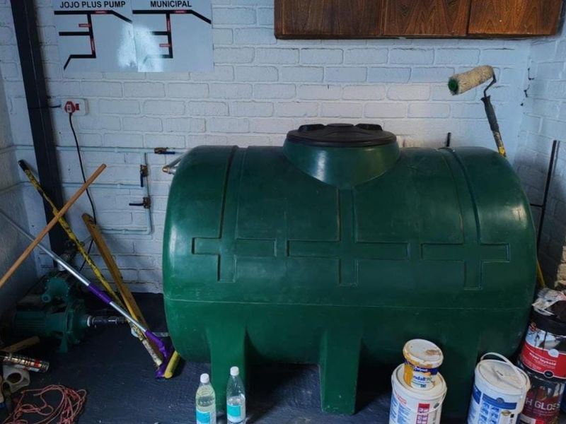 Utility area featuring a large green water storage tank, integrated water pump system, and exposed plumbing infrastructure against a painted brick wall.