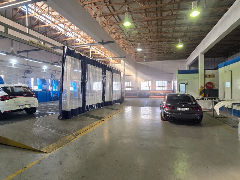 A spacious commercial automotive workshop featuring industrial lighting, exposed roof trusses, concrete flooring, and dedicated vehicle preparation and spray booth areas.