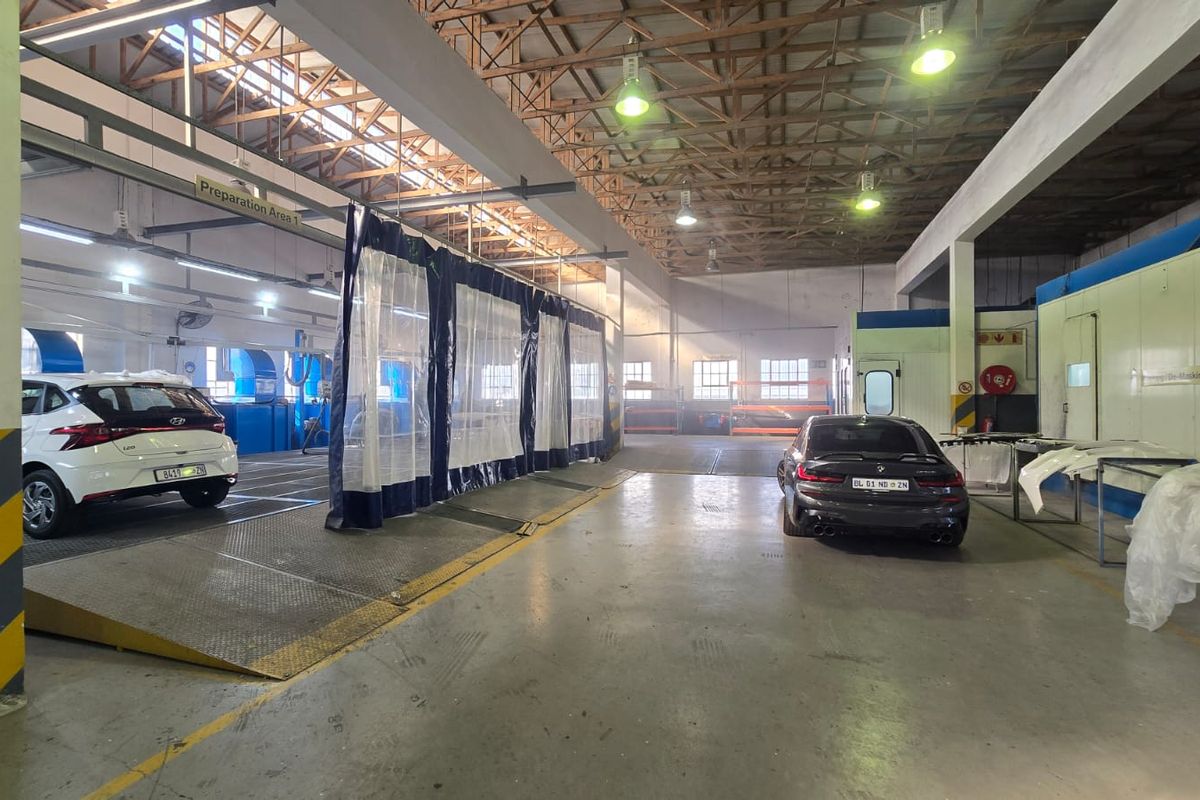 A spacious commercial automotive workshop featuring industrial lighting, exposed roof trusses, concrete flooring, and dedicated vehicle preparation and spray booth areas.