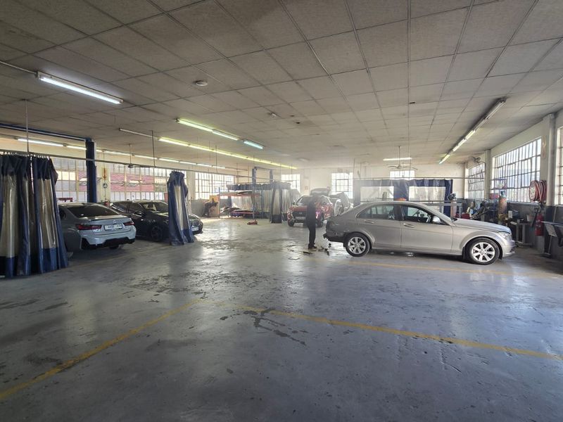 Spacious commercial automotive workshop featuring industrial concrete flooring, multiple service bays, fluorescent lighting, and large windows providing ample natural light.