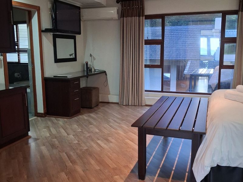 A well-maintained hotel bedroom featuring laminate flooring, a built-in desk, split-system air conditioning, and large windows with views of the exterior.