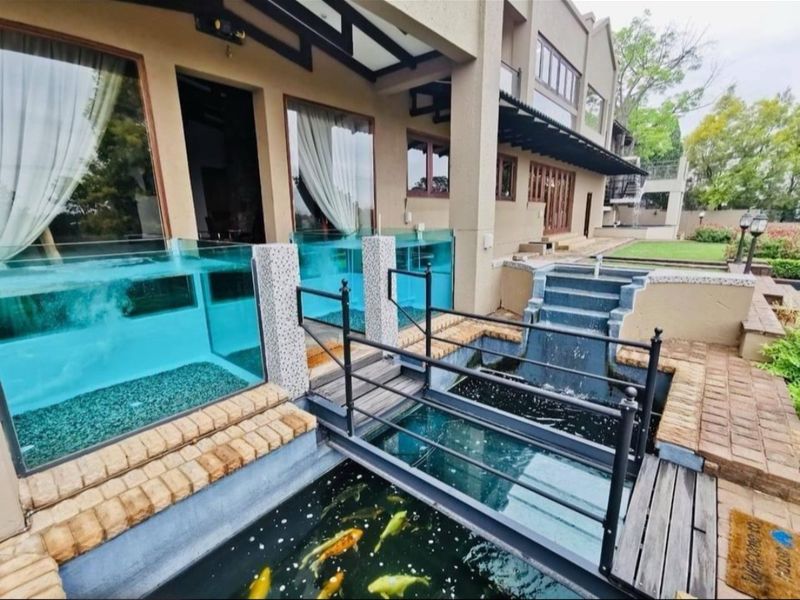A beautifully landscaped rear exterior featuring a custom koi pond, a glass-walled pool, and a pedestrian bridge leading to the covered patio area.