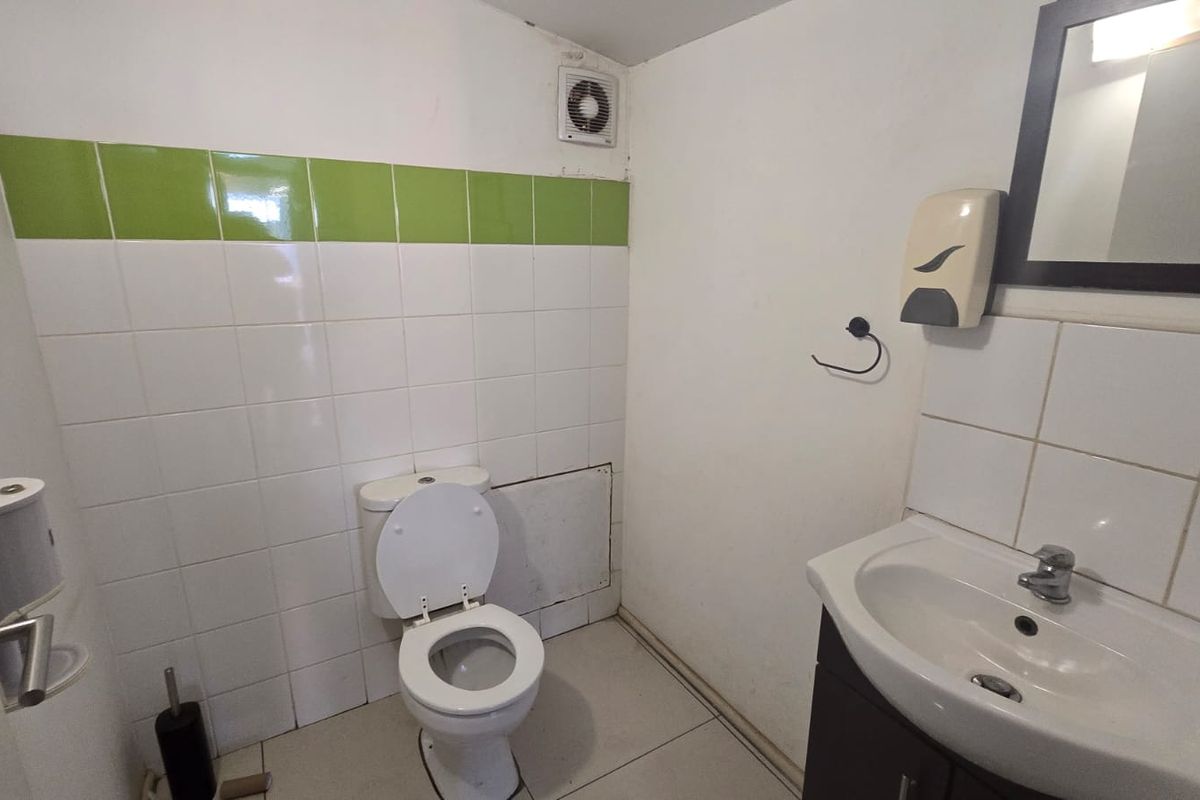 A functional commercial bathroom featuring a standard toilet, vanity basin, and partial wall tiling with an extractor fan.