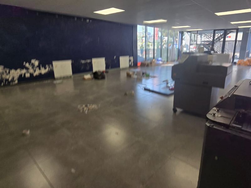 Spacious open-plan commercial area featuring large glass windows, tiled flooring, and recessed lighting, currently in need of cosmetic wall repairs.