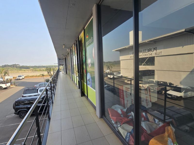 An elevated exterior walkway featuring tiled flooring, metal safety railings, and large glass storefront windows overlooking a commercial parking area.
