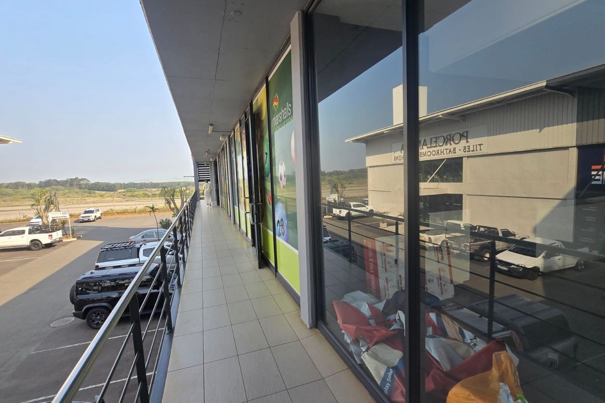 An elevated exterior walkway featuring tiled flooring, metal safety railings, and large glass storefront windows overlooking a commercial parking area.