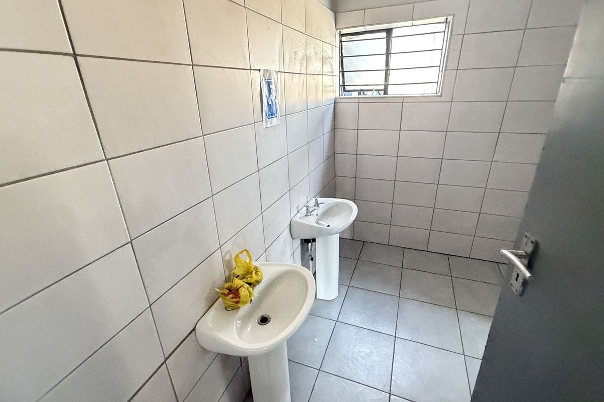 A functional industrial-style washroom featuring dual pedestal basins, fully tiled walls and floors, and a window with security bars.