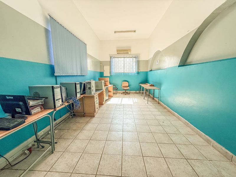 A functional office space featuring ceramic tile flooring, a wall-mounted air conditioning unit, and security-barred windows with vertical blinds.