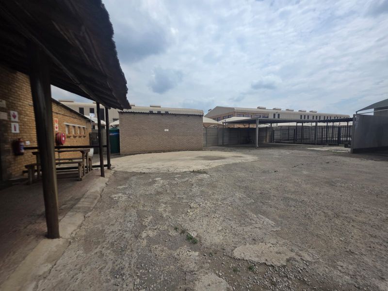 Rear exterior area of the restaurant featuring a covered patio section, brick structures, and a large paved parking or utility yard.