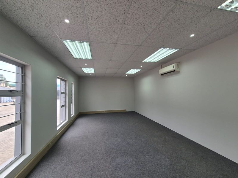 A clean, empty office space featuring a suspended ceiling, recessed lighting, wall-mounted air conditioning, and carpeted flooring with large windows.