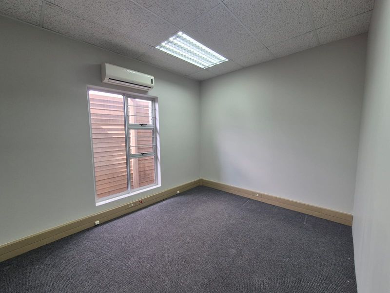 A clean, empty office space featuring grey carpeting, a suspended ceiling with recessed lighting, and a wall-mounted air conditioning unit.