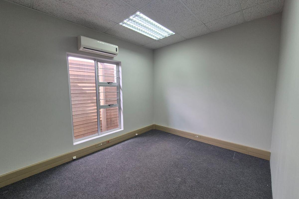 A clean, empty office space featuring grey carpeting, a suspended ceiling with recessed lighting, and a wall-mounted air conditioning unit.