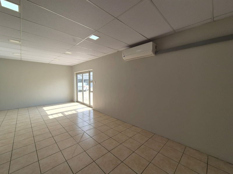 A bright, empty office space featuring tiled flooring, a suspended ceiling with recessed lighting, and a wall-mounted air conditioning unit.