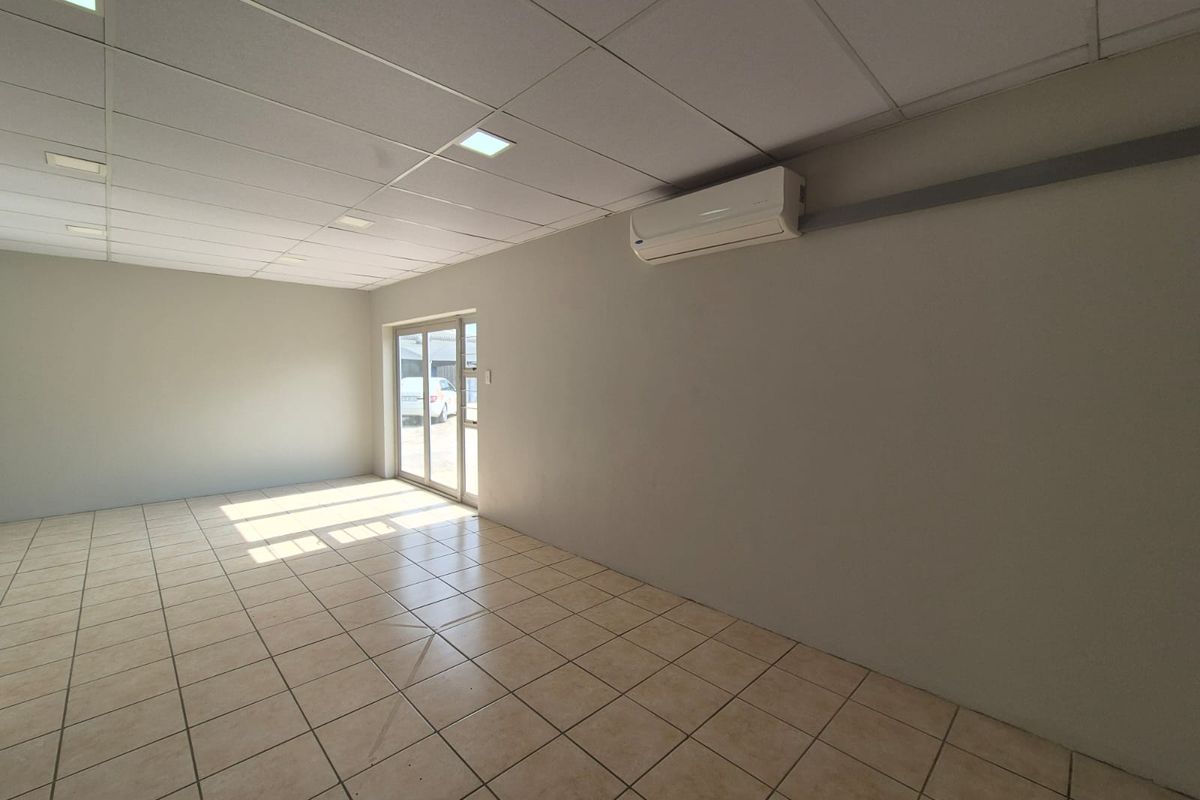 A bright, empty office space featuring tiled flooring, a suspended ceiling with recessed lighting, and a wall-mounted air conditioning unit.