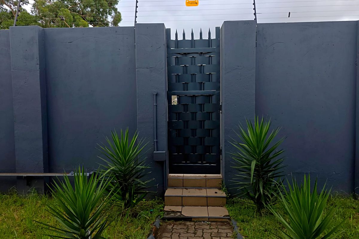 Secure rear access point featuring a high perimeter wall with electric fencing, a metal security gate, and a paved walkway leading to tiled steps.