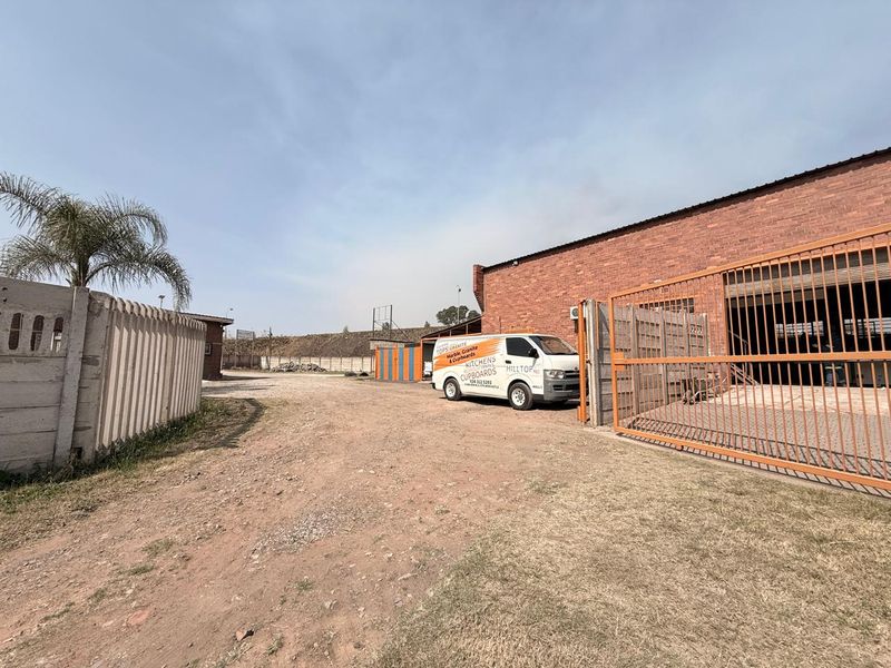 Industrial property exterior featuring a brick warehouse building, secure metal gate access, and an unpaved yard area.