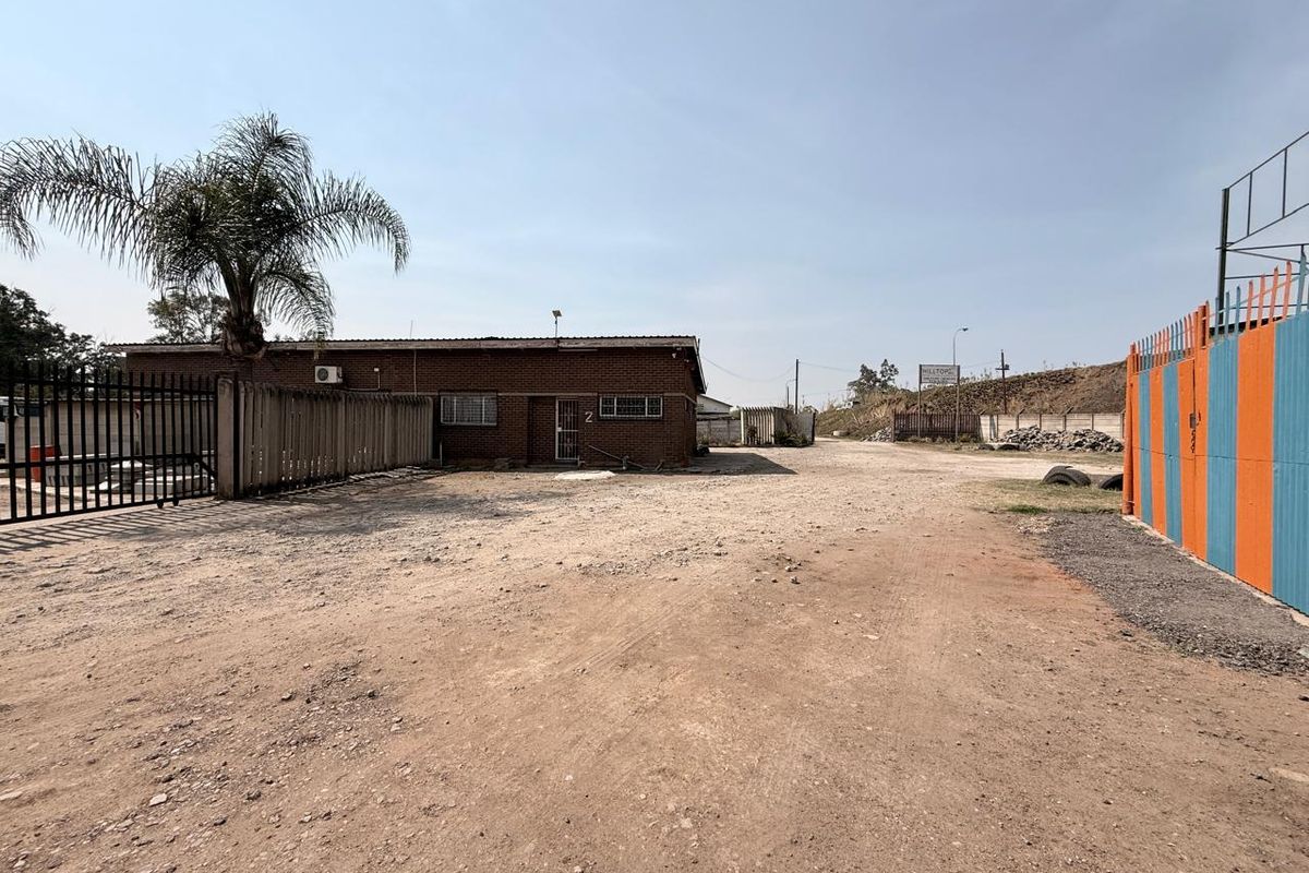 Front exterior view of an industrial property featuring a brick building, security fencing, and an unpaved yard area.