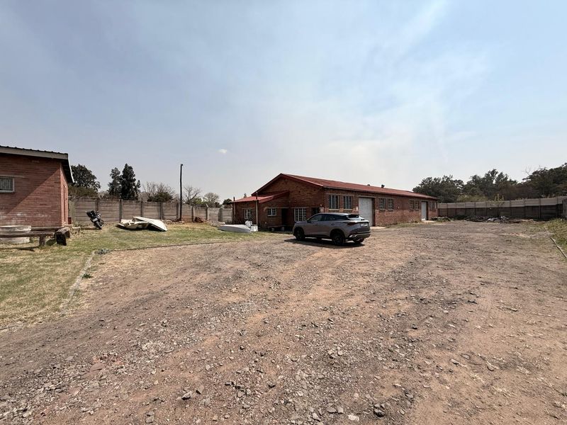 Spacious industrial property exterior featuring a large gravel yard, brick buildings with metal roofing, and secure perimeter walling.