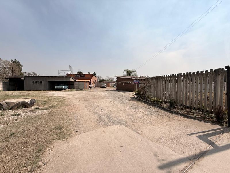 Industrial property entrance featuring a gravel driveway, perimeter concrete wall, and multiple brick warehouse structures with corrugated metal roofing.