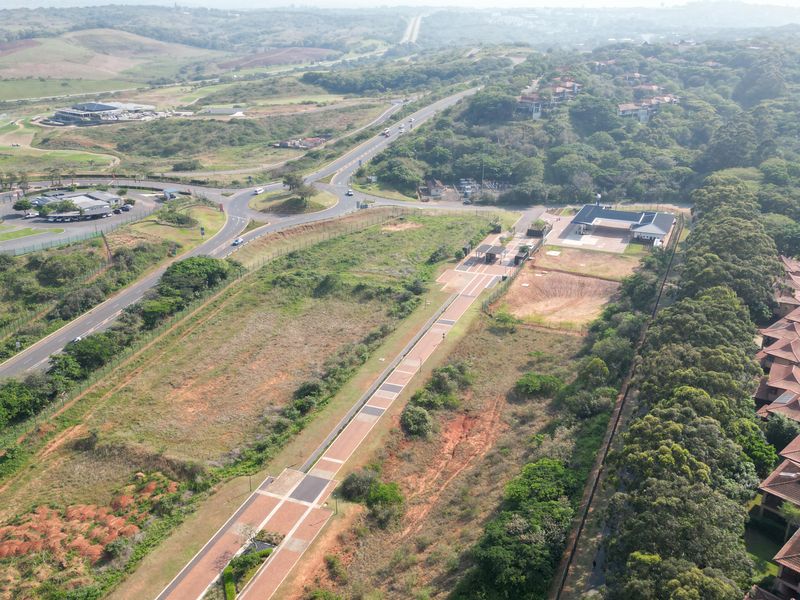 Aerial view of a commercial vacant land plot featuring a paved access road, security gatehouse, and established perimeter fencing in a developed area.