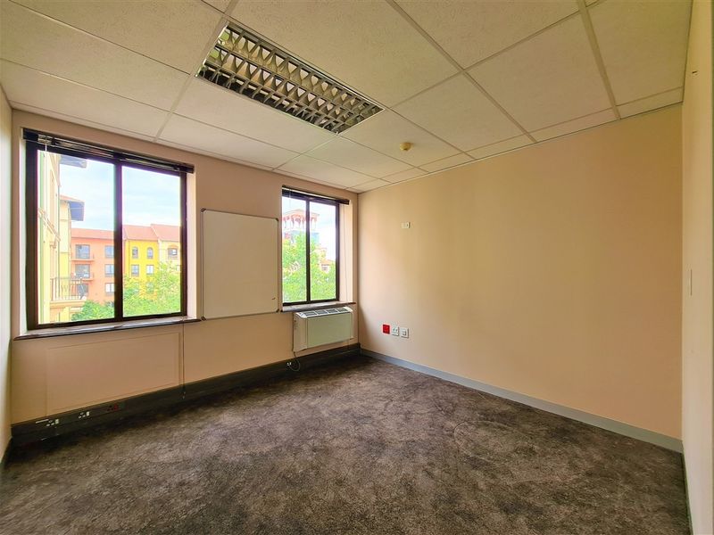 A vacant office space featuring a suspended ceiling, recessed lighting, wall-mounted HVAC unit, and carpeted flooring with large windows providing natural light.