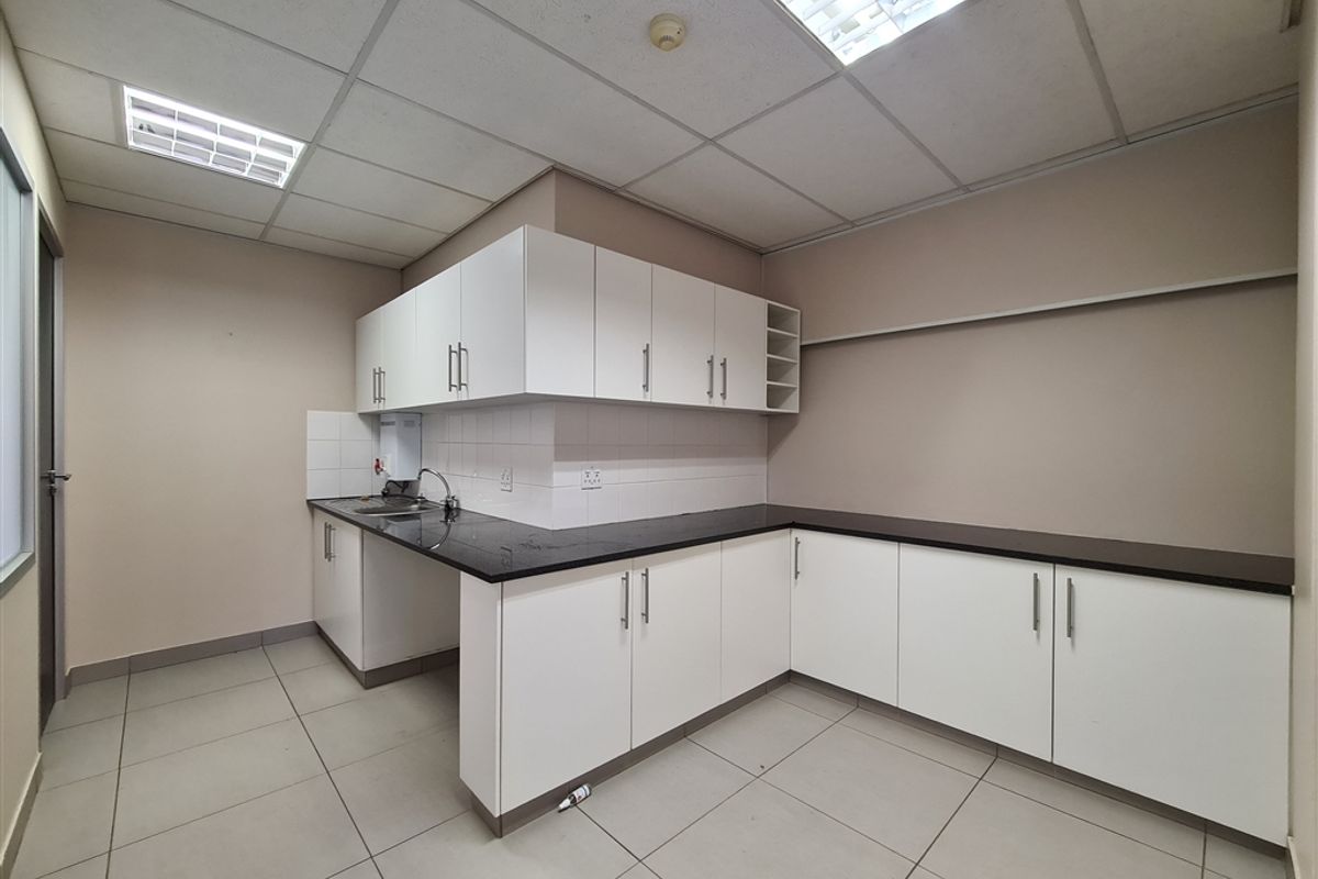A clean, functional office kitchenette featuring white cabinetry, dark laminate countertops, a stainless steel sink, and neutral ceramic floor tiles.